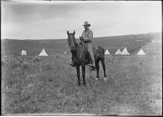 A black-and-white image of a cowboy mounted on a horse standing in a field with several tipis and tents in the background.