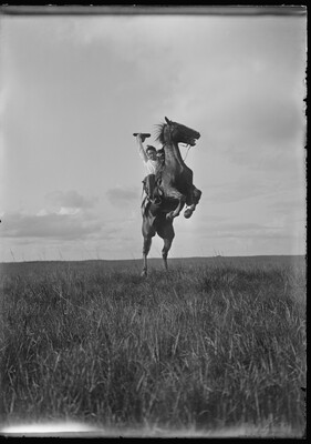 A black-and-white image of a White man on a rearing horse holding his hat in one hand above his head.