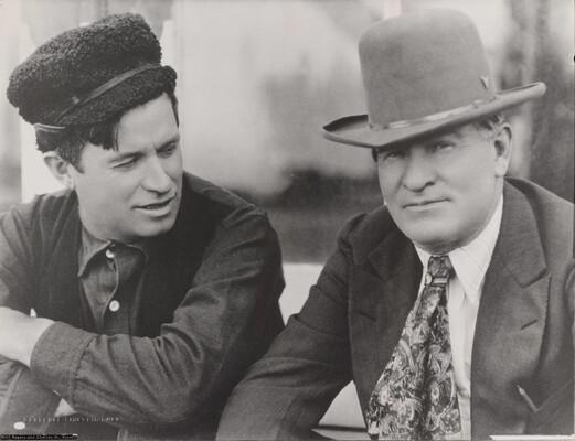 A black-and-white photograph of two White men: the man on the left wearing a fuzzy hat, dark vest, and button-down shirt; the man on the right wearing a cowboy hat with a jacket and tie.