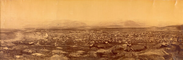 A sepia-toned panoramic photograph taken from high up of a small town in a barren, rocky landscape with mountains in the distance.
