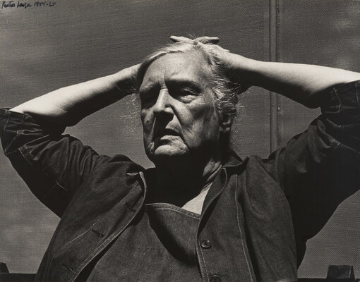 A black-and-white photograph of an older White woman, tired expression on her face, wearing a denim jacket, her hands folded on top of her head.