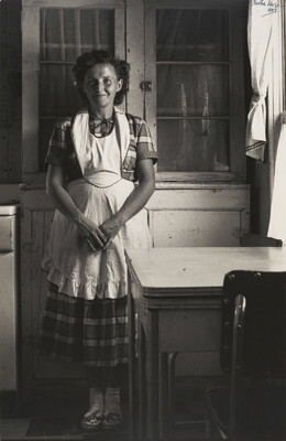 A black-and-white photograph of a short-haired, smiling White woman wearing a plaid dress under an apron, standing in a kitchen in front of empty cabinets.