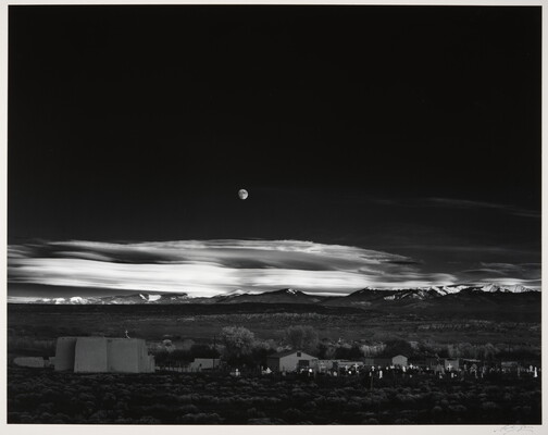 A black-and-white landscape photograph of a town with mountains in the background and the moon rising above the clouds in the night sky.