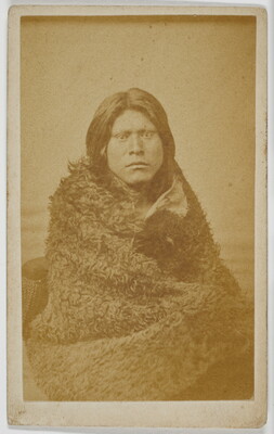A sepia-toned studio photograph of a seated Indigenous person wrapped in a fur blanket.