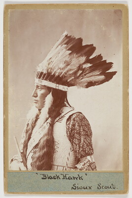 A black-and-white photograph of an Indigenous person with long dark hair, wearing a feathered headdress, a light colored vest and standing in profile.