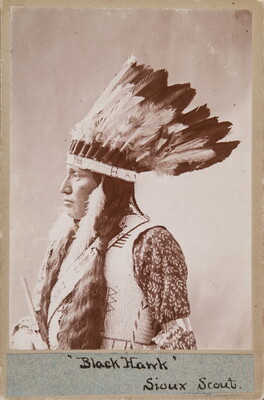 A black-and-white photograph of an Indigenous person with long dark hair, wearing a feathered headdress, a light colored vest and standing in profile.