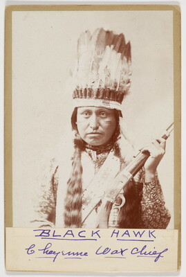 A black-and-white photograph of an Indigenous person with long dark hair, wearing a feathered headdress, a light colored vest and holding a rifle.
