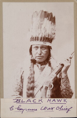 A black-and-white photograph of an Indigenous person with long dark hair, wearing a feathered headdress, a light colored vest and holding a rifle.