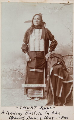 A sepia-toned full-body portrait of an Indigenous man in cultural dress, including a breastplate.