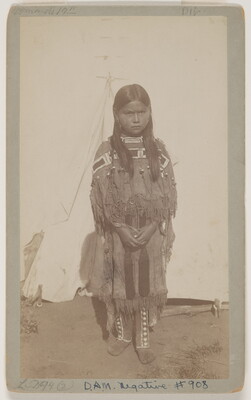 A sepia-toned photograph of a young Native American girl in traditional clothing standing outside.