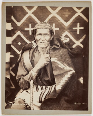 A sepia-toned photograph of an Indigenous person wearing a cloth turban, seated in front of woven blankets with another blanket wrapped around their shoulders.