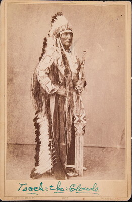 A sepia-toned portrait photograph of an Indigenous chief standing in traditional regalia, including a full-feathered headdress.