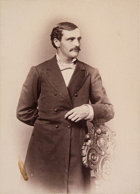 A sepia-toned portrait of a White man wearing a long frock coat standing posed with his arm on the back of a chair.