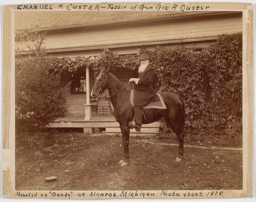 A sepia-toned photograph of a bearded White man seated on a horse in front of a house covered in vines.