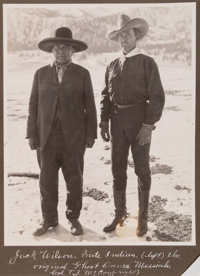 A black-and-white photograph of an Indigenous man in a suit and wide-brimmed hat standing next to a White man in dark clothes and a cowboy hat.