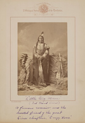 A black-and-white formal portrait photograph of an Indigenous man in traditional clothing surrounded by a headdress and several Indigenous objects.