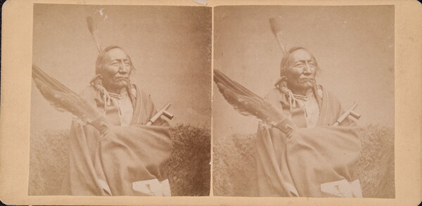 A sepia-toned stereograph of an Indigenous man holding a group of large feathers and a pipe.