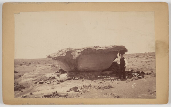A sepia-toned photograph of a man standing next to a bowl-shaped rock formation in the desert.