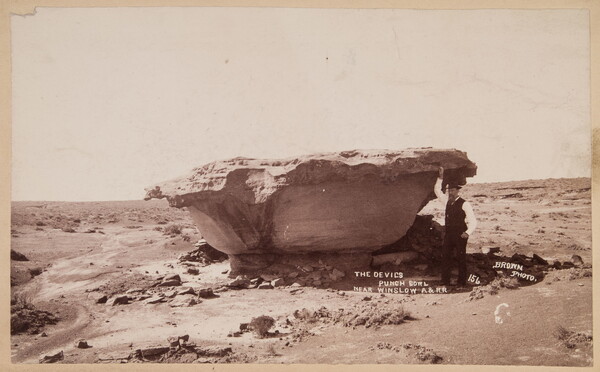 A sepia-toned photograph of a man standing next to a bowl-shaped rock formation in the desert.