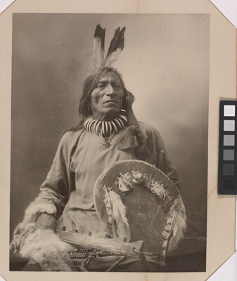 A black-and-white formal portrait photograph of a seated Indigenous man in traditional clothing holding a shield.