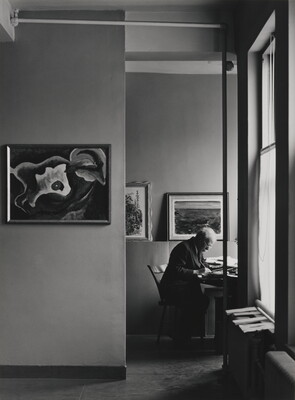 A black-and-white photograph of an older White man working at a desk in a room lit by sunlight through windows, artworks hanging on the walls.