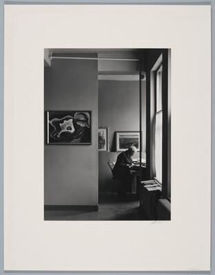 A black-and-white photograph of an older White man working at a desk in a room lit by sunlight through windows, artworks hanging on the walls.