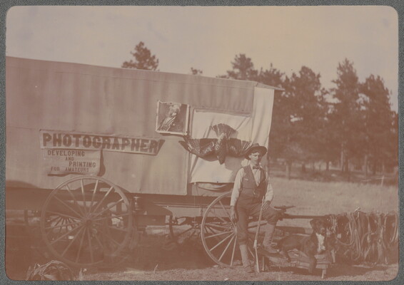 A black-and-white photograph of a White man and a dog posing in front of a covered wagon adorned with a sign that reads, "Photographer."