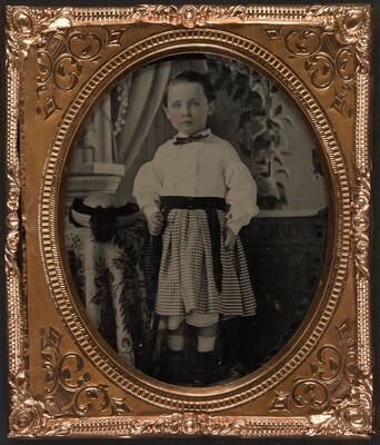 A black-and-white photograph in a decorative gold frame of a White child wearing a white shirt and plaid skirt, standing beside a table.