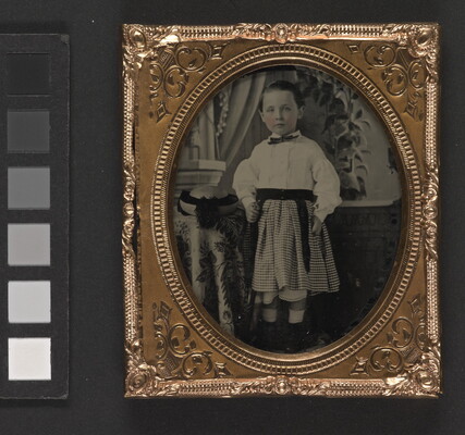 A black-and-white photograph in a decorative gold frame of a White child wearing a white shirt and plaid skirt, standing beside a table.
