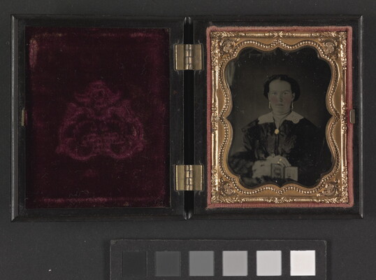 A black-and-white photograph in a decorative gold frame, open to show the red velvet, of a White woman in dark dress with a white collar and brooch at the neck holding a cased photograph on her lap.
