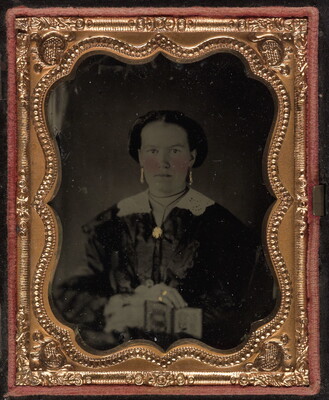 A black-and-white photograph in a decorative gold frame of a White woman in dark dress with a white collar and brooch at the neck holding a cased photograph on her lap.