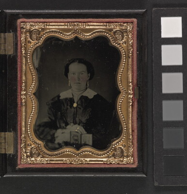 A black-and-white photograph in a decorative gold frame of a White woman in dark dress with a white collar and brooch at the neck holding a cased photograph on her lap.