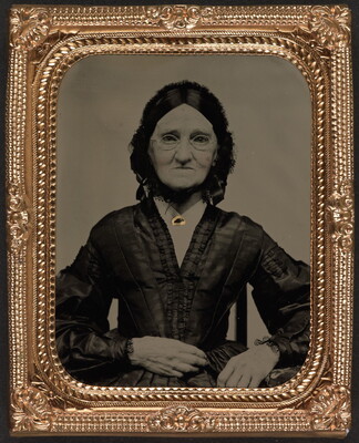 A black-and-white photograph in a decorative gold frame of an elderly White woman in spectacles wearing a dark Victorian-style dress.