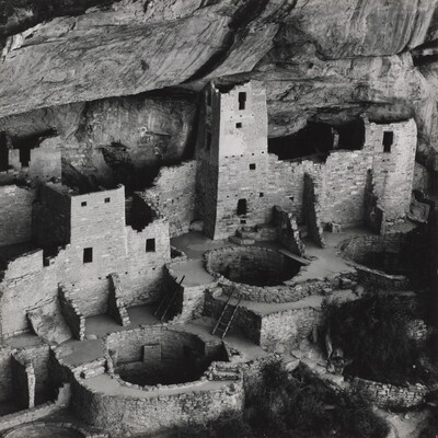 A black-and-white photograph of ruined stone buildings, some with towers, and several underground circular rooms all under the overhang of a cliff.