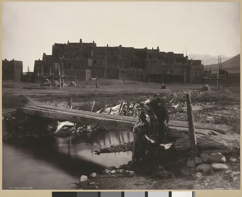 A black-and-white photograph of two Indigenous people by a footbridge over a creek with a large adobe pueblo in the background.