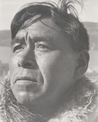 A black-and-white photograph of a close-up of a middle-aged Indigenous man with windblown hair and fur at his neck.