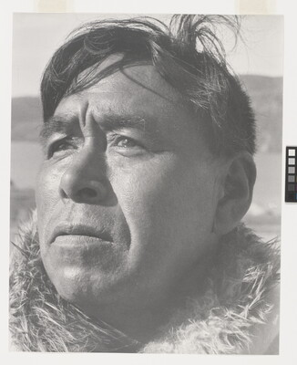 A black-and-white photograph of a close-up of a middle-aged Indigenous man with windblown hair and fur at his neck.