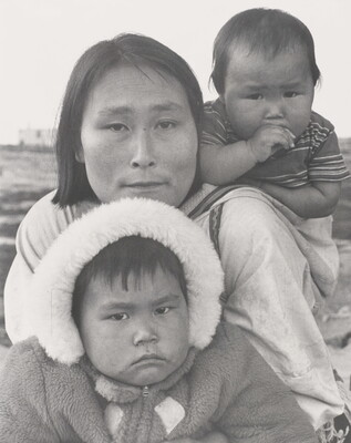 A black-and-white photograph of an Indigenous woman with a baby on her back and another child wearing a hooded coat in front of her.
