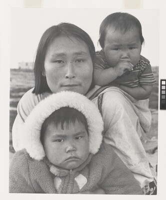 A black-and-white photograph of an Indigenous woman with a baby on her back and another child wearing a hooded coat in front of her.