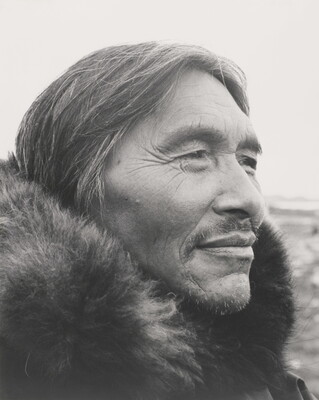 A black-and-white photograph of a close-up of a middle-aged Indigenous man, salt-and-pepper hair and wrinkles around his eyes, a mustache and a fur at his neck.