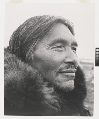 A black-and-white photograph of a close-up of a middle-aged Indigenous man, salt-and-pepper hair and wrinkles around his eyes, a mustache and a fur at his neck.