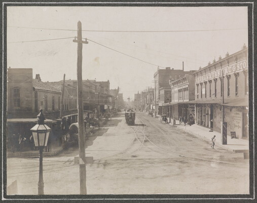 A black-and-white photograph of a western-style town with a trolley on tracks in the center of an unpaved street.