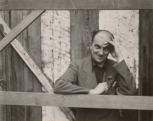 A black-and-white photograph of a bald, White man standing next to a wood structure, wearing a suit jacket and resting his head in his hand.