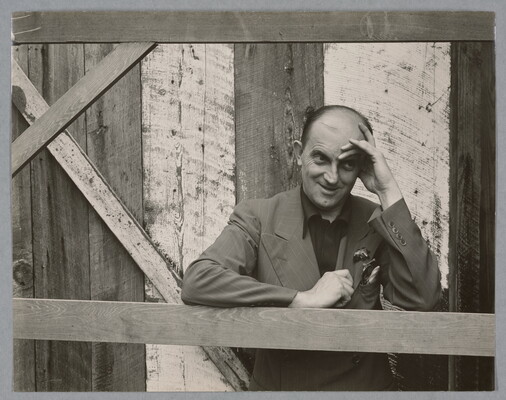 A black-and-white photograph of a bald, White man standing next to a wood structure, wearing a suit jacket and resting his head in his hand.