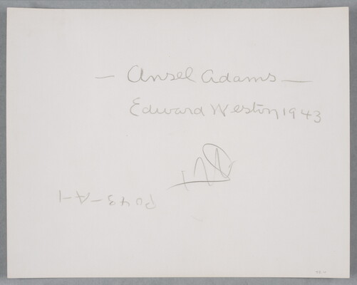 A black-and-white photograph of a bald, White man standing next to a wood structure, wearing a suit jacket and resting his head in his hand.