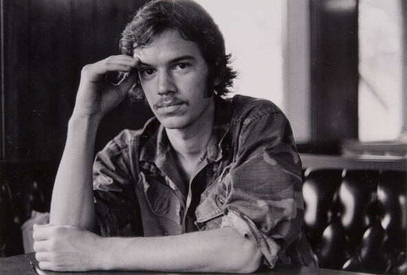 A black-and-white photograph of young White man wearing a button-down shirt with the sleeves rolled up, one arm resting on the table in front of him, the other raised to his temple.
