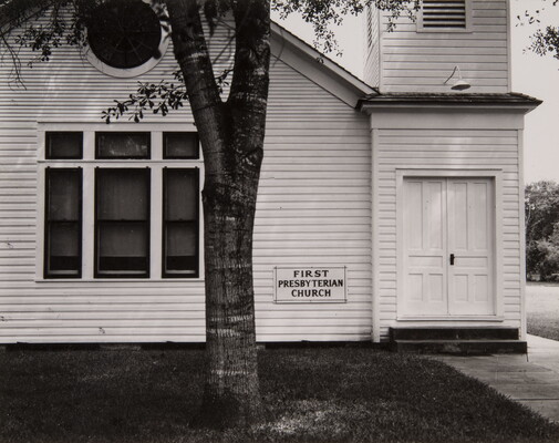 A black-and-white photograph of a white clapboard church with a sign that reads "First Presbyterian Church" by the door and a tree in the foreground.