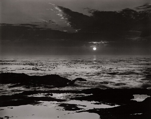A black-and-white photograph of waves crashing on a beach under a cloudy sky and the sun low on the horizon.