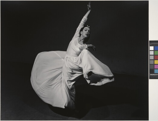 A black-and-white studio photograph of a woman wearing a long white dress that swirls as she dances.