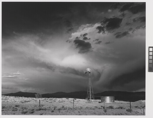 A black-and-white photograph of a windmill, black hills in the distance, under a stormy sky.